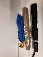 Top view of three closed umbrellas: blue with bird head handle, black with plastic handle, and tan umbrella in packaging.