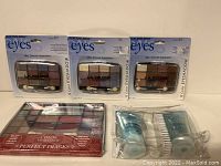 Three unopened L.A. Colors 12-color eyeshadow palettes with neutral and purple tones in sealed plastic packaging, located at the back, and several other beauty items including a lip palette and packaged eyelash curlers in front.