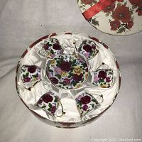 Top view of the porcelain tea set including six tea cups arranged around a central saucer inside the round box lined with white satin fabric.