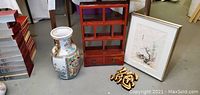Four items on floor: a red lacquered wooden Asian tea shelf with two small drawers, a framed signed Chinese botanical painting, a tall multicolored Chinese porcelain vase, and a gold-toned enameled resin Chinese character wall art.