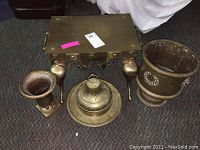 Photo showing brass seat with ornately styled legs and handles, two brass urns of different sizes, and a brass incense pot with lid and tray on carpeted floor.