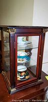 Photo of tall wooden and glass curio box with three shelves visible, containing porcelain teacups and saucers, showing box and contents