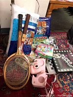 Photo of lot showing two large bags of polyester fiberfill, various board games including Monopoly Token Madness and Disco Steppin, a tie-dye kit, two wooden tennis rackets, and a pink Instax camera with case.