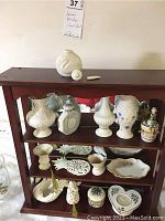 Full frontal view of dark wooden three-shelf curio cabinet with porcelain objects arranged on shelves including vases, trinket boxes, plates, and candle holder.