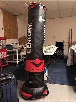 Full view of tall Century Versys punching bag in black and red, standing upright surrounded by miscellaneous items in background.