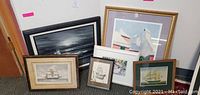 Photo showing seven framed artworks arranged on the floor leaning against a wall, including maritime and coastal scene prints and paintings.
