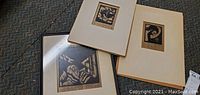 Three unframed woodblock engraving prints lying on a carpet. Two prints are mounted on cream color mat boards, one is framed in black.