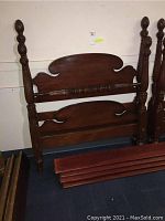 Front view of wooden twin bed headboard, footboard, and side rails stacked against wall showing carved posts and scratches