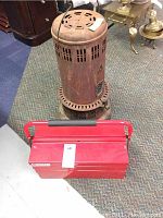 Photo of rusty cylindrical vintage oil heater with ventilation and black base. Red metal toolbox with black cushioned handle in front.