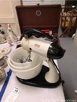 Vintage Sunbeam Mixmaster stand mixer with white mixing bowls and beaters placed on a table with a chest of flatware in the background