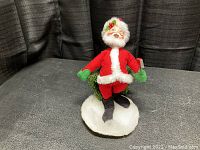 Annalee Santa doll dressed in red with green gloves and black boots on a snow base holding a small green Christmas tree.