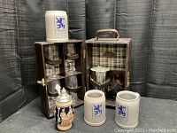 Wooden case containing stemmed glasses, two Lowenbrau ceramic mugs, German stein, and metal ice bucket with tongs arranged on black cloth background