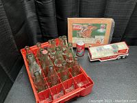 Lot showing 11 vintage Coca-Cola glass bottles in a red crate alongside a Coca-Cola can, diecast truck, and vintage ad print