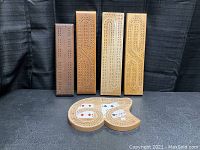 Five wooden cribbage boards standing vertically, showing different wood colors and hole patterns. The uniquely shaped board laid in front highlights its card holder design and printed cards.