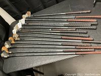 Full set of 12 golf clubs with a mix of wood and iron heads laid out side by side on a table, showing grips and shafts.