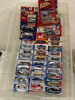 Photo showing diecast cars from various brands including Hot Wheels Classics, Johnny Lightning, and Winners Circle, all in blister card packaging organized in a plastic storage bin.