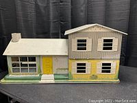 Front exterior view of the Marx tin bilevel doll house showing white shingled roof, yellow front door, window shutters, and lithographed siding and brick walls.