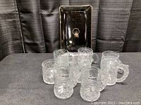 Photo showing six clear glass mugs with embossed Batman faces and logos along with a rectangular glass tray with gold trim.