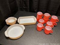 Full set of peach luster bowls, rectangular and round dishes with 10 orange stackable mugs in front on black surface with black curtain backdrop.