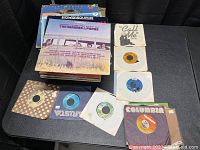 Stack of vinyl record albums and several 45-RPM singles laid out showing front album covers and singles in paper sleeves.