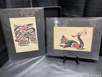 Two framed and matted Native style art prints side by side with plastic protective covers. One print on the left depicts a heavily detailed bird design and the other on the right shows a long-necked bird.