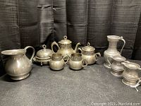 Photo showing the full collection of silver plated tea sets and pewter pitchers arranged on a surface with black backdrop.