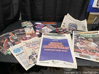 Photo showing a mix of sports newspapers, signed and unsigned card packs, photos, and memorabilia sticker on a table with a black curtain background.