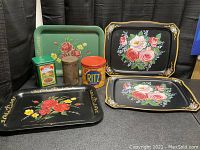 Photo showing three black metal trays with floral painted roses in varying sizes and shapes, two trays have handles, one does not. Three tin containers are placed in front.