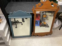 Front view of two vintage wooden framed mirrors, one blue framed with scalloped top and one natural wood frame with ornate carved top.