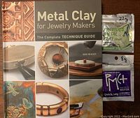 Front cover of the Metal Clay book with silver PMC+ and Standard Silver PMC clay packets next to it