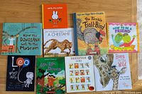 Top-down photo showing all 10 children's books arranged on a wooden floor, titles clearly visible including dinosaur, artist, cheetah, troll bird, friendship and more.