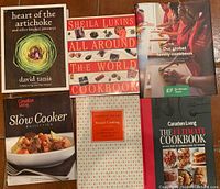 Top view of six cookbooks laid out on a brown surface showing front covers including Heart of the Artichoke, Sheila Lukins All Around the World, Our Global Family Cookbook, Canadian Living Slow Cooker Collection, Mastering the Art of French Cooking, and Ultimate Cookbook by Canadian Living.