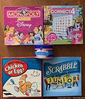 Four children's board games arranged on wooden floor: Monopoly Junior Disney, My Little Pony Connect 4, Chicken or Egg?, Scrabble Junior.