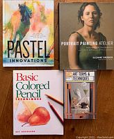 Four art instruction books arranged on wooden surface: Pastel Innovations, Portrait Painting Atelier, Basic Colored Pencil Techniques, and Art Terms & Techniques.