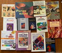 Overview photo showing 13 arts and crafts instructional books arranged on a wooden floor. Titles include Fimo, Papier Mâché, Stained Glass, Watercolor, Marker Rendering, Textile Arts, Illustration, Collage Techniques, Dye Painting, and pigment theory.
