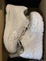 Side view of both white leather running shoes side by side inside the box showing outsole, stitching, and texture.