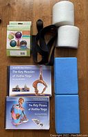 View of two yoga anatomy books, two blue blocks, black strap, two white foam rollers, and boxed massage balls and foot roller.