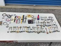 Full overview of all items laid out on white table including numerous collector spoons arranged in rows, several wristwatches, bracelets, small boxes, and pins