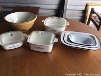 Photo showing all items in the lot: three Corning Ware Plus casserole dishes with lids, large mixing bowl, and three white oval nested trays with blue rims.