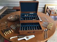 Photo showing wooden presentation case open with assorted metal flatware inside, additional knives and forks laid around case on table surface with varying handle styles and colors.