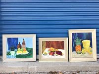 Three framed still life paintings displayed outdoors on concrete with blue corrugated metal background