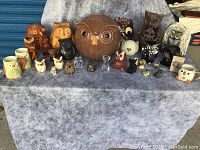 Front view showing full collection of owl figurines and mugs on table with grey cloth background. Shows variety in size, color, and materials.