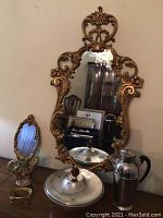 Photo showing large ornate gold-tone mirror on wall along with smaller mirror, ceramic pitcher, covered dish, and silver thermal carafe on tabletop