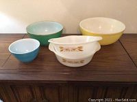 Photo showing the full set of four vintage mixing bowls including two white FireKing wheat pattern bowls, a medium blue-green bowl, small blue bowl, and large yellow bowl placed on a table.