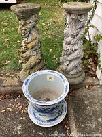 Two cement bird bath pedestals with detailed textured carvings next to a blue and white ceramic planter with underplate, set outside on patio.