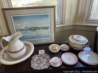 Photo showing framed seascape print by Richard Grosvenor, porcelain pitcher and washbowl, cups with saucers, chintz plate, and various plates on a dark wooden surface near window