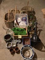 Photo showing an arrangement of wicker baskets, plastic gardening containers, small plant pots, and a black watering can on a concrete floor.
