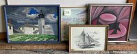 Four framed artworks grouped on a wooden surface, including lighthouse and buildings oil painting, beach scene oil painting, sailing ship drawing, and pink ladies' hat painting.