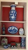 Front view of shelving unit containing large blue and white oriental ceramic vase on top shelf, colorful folk figurine, blue and white oval platter, oval vase on middle shelf, and ceramic planter, jar, and baoding balls on bottom shelf.