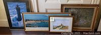 Group photo of six framed artworks including lighthouse, harbor, beach, and family scenes stacked and leaning against wall.